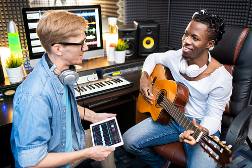 Young man with digital tablet talking to happy colleague with guitar while both sitting at workplace in sound recording studio
