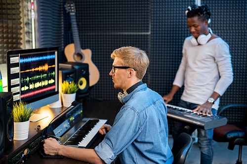 Serious young man in denim shirt sitting in front of computer screen and working with sound waveforms while another guy playing pianoboard
