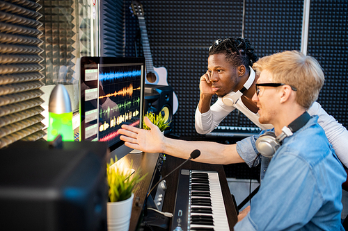 Young African musician looking at sound waveforms on computer screen while his colleague pointing at one of them in studio