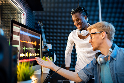 Young musician or producer showing African man group of sound waveforms on computer screen while working in team