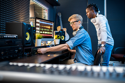 Young casual man adding volume while recording music with his African colleague in contemporary studio