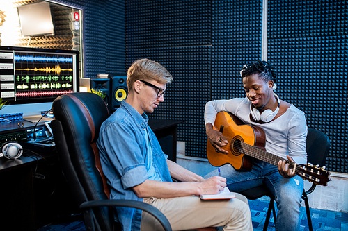 Two young multicultural men playing guitar and making notes in notepad while making song in sound recording studio