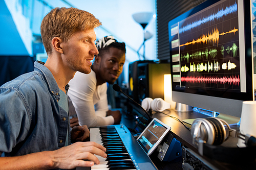 Young man with pianoboard sitting in front of computer screen and looking at sound waveforms with colleague on background