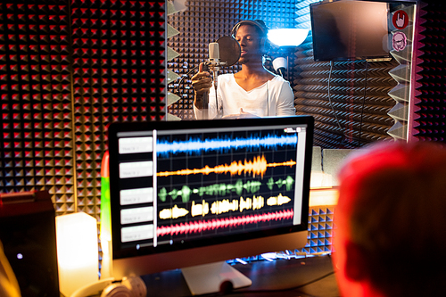 Young African-american man with headphones singing in microphone while producer recording song in front of computer screen