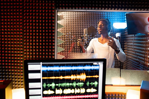 Young man of African ethnicity singing in microphone in sound recording studio while his performance is being recorded