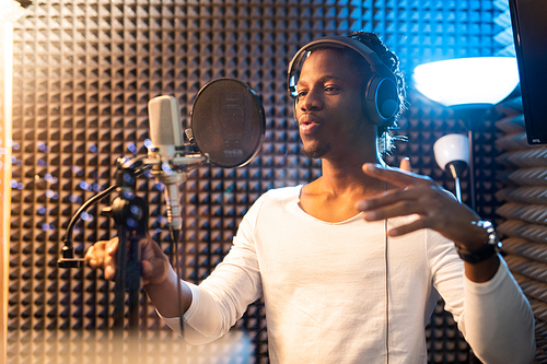 Young African man in white t-shirt and headphones standing by microphone and performing songs in studio of sound recording