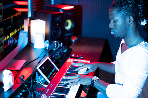 Young serious African-american man making new music while sitting in studio in front of pianoboard and pressing keys