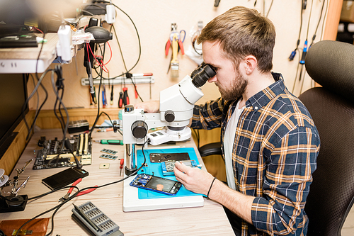 Young bearded repairman sitting by table in front of microscope to see tiny details of broken gadgets in workshop