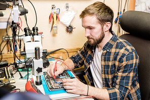 Young serious mechanic with two soldering-irons repairing demounted touchpad by workplace in workshop