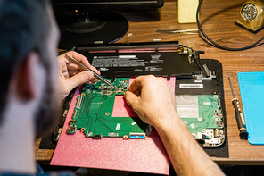 Tweezers in hands of repairman over demounted gadget during work with tiny details by table in workshop