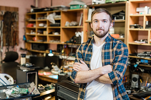 Young professional repairman of broken gadgets crossing arms by chest while standing in front of camera in his workshop