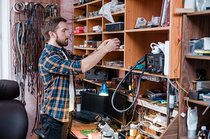 Young repairman in casualwear standing by workplace and taking working supplies from one of shelves