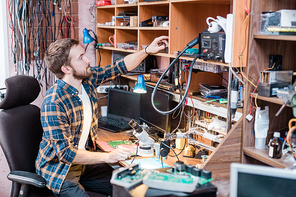 Young professional repairman sitting in armchair by table while taking working supplies from one of shelves