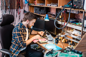 Young professional mechanic with small steel tweezers repairing demounted touchpad or other gadget in his workshop