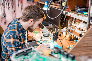 Professional gadget repairman looking in microscope by workplace while trying to find problem of smartphone or tablet