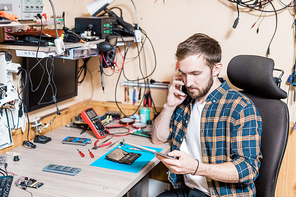 Young professional master of gadget repair service talking to client on smartphone while looking through online requests in touchpad