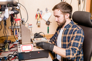 Bearded repairman in protective gloves sitting by workplace in front of equipment for gadget repair and using it