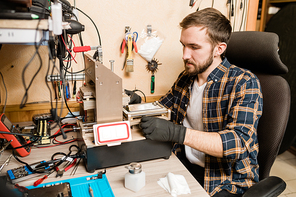 Bearded repairman in protective gloves sitting by working equipment while trying to find out trouble of broken smartphone