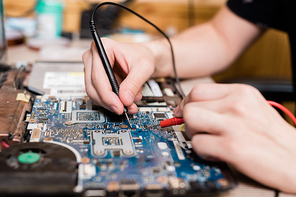 Hands of young master of gadget repair service using two small soldering-irons while repairing demounted laptop