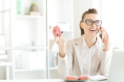 Cheerful young businesswoman sitting by desk in front of laptop, eating tasty donuts and talking to friend by smartphone