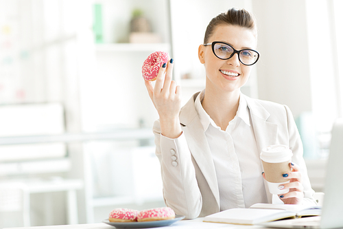 Young office manager with donut and coffee  by her desk during break for some snack