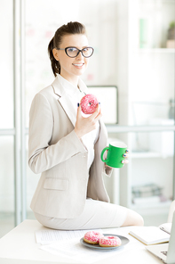 Happy young female with donut and glass of hot coffee sitting on her desk and having snack in office