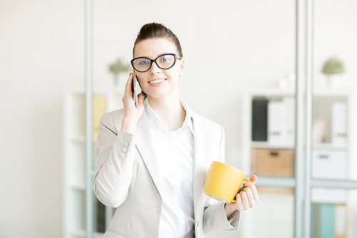 Successful office manager with yellow mug speaking by smartphone before coffee break
