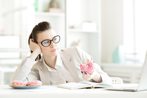 young businesswoman eating tasty donuts and watching something in the net while sitting by desk in front of laptop