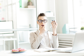 Elegant businesswoman applying lipstick or gloss on her lips by workplace in office