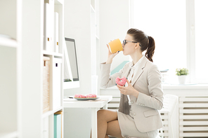 Young hungry businesswoman drinking coffee from yellow mug and eating fresh donuts by her workplace