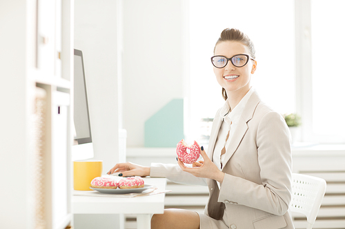 Young smiling businesswoman with donut  during lunch break in front of computer monitor