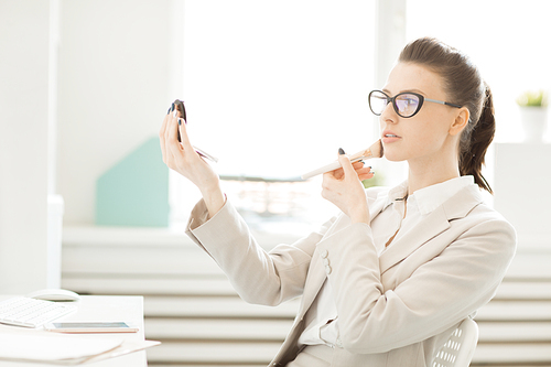 Young pretty businesswoman looking in compact powder mirror while applying blush on her cheeks