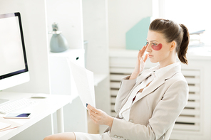 Modern businesswoman with under-eye patches on face sitting by workplace and reading contracts or documents