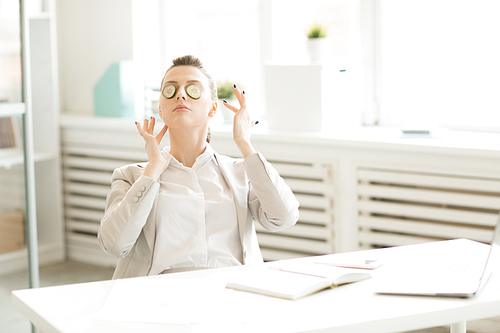 Young businesswoman sitting by desk in office with slices of fresh cucumber on her eyes during break