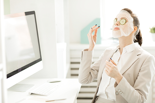 Young female employee in suit having cotton facial mask and slices of fresh cucumbers on eyes while sitting by workplace with computer monitor