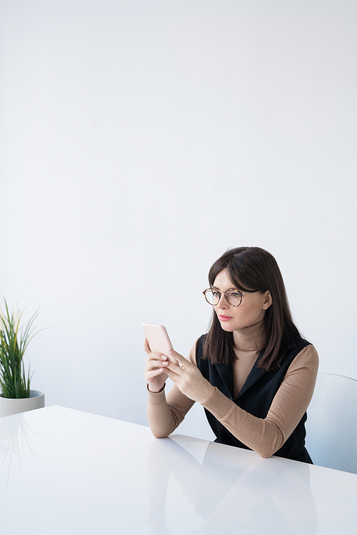 Pretty young businesswoman in smart casual scrolling in smartphone while sitting by desk against white wall in office