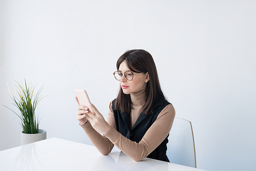 Young brunette businesswoman in smart casual scrolling in smartphone while sitting by desk in office