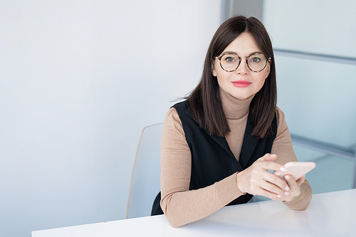 Pretty young businesswoman in smart casual and eyeglasses using smartphone while sitting by desk against white wall in office