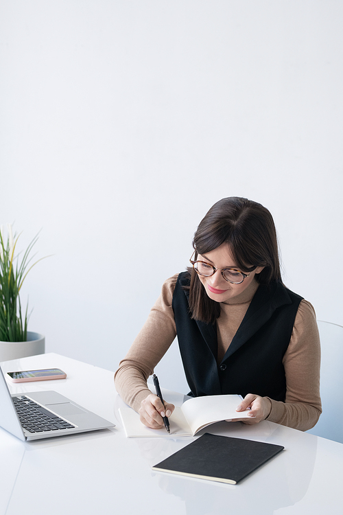 Young successful businesswoman writing down plan of working day while sitting by desk in front of laptop