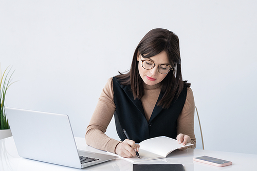Young elegant teacher or businesswoman making working notes in copybook while sitting in front of laptop