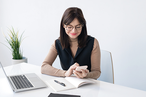 Young teacher or businesswoman with smartphone over open copybook looking for contact while sitting by desk in front of laptop