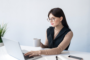 Young serious businesswoman with mug analyzing online financial data while sitting in front of laptop in office