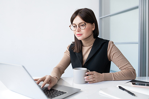 Young contemporary businesswoman with drink analyzing online financial data in front of laptop in office