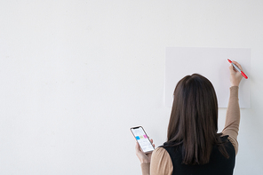 Back view of young businesswoman or teacher with smartphone and highlighter standing by whiteboard