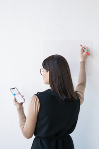 Rear view of elegant businesswoman or coach with smartphone making presentation on whiteboard