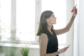 Elegant coach with smartphone writing down on whiteboard while making presentation for students