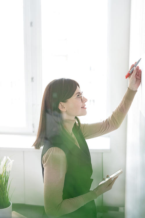 Young successful businesswoman with smartphone preparing presentation on whiteboard in office