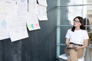 Pretty young student or businesswoman sitting on chair and making notes while looking at papers on blackboard