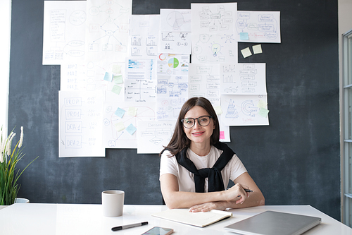 Young successful female economist sitting by desk on background of blackboard with papers and working