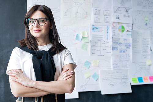 Pretty young businesswoman in eyeglasses crossing arms by chest while standing in front of camera in office